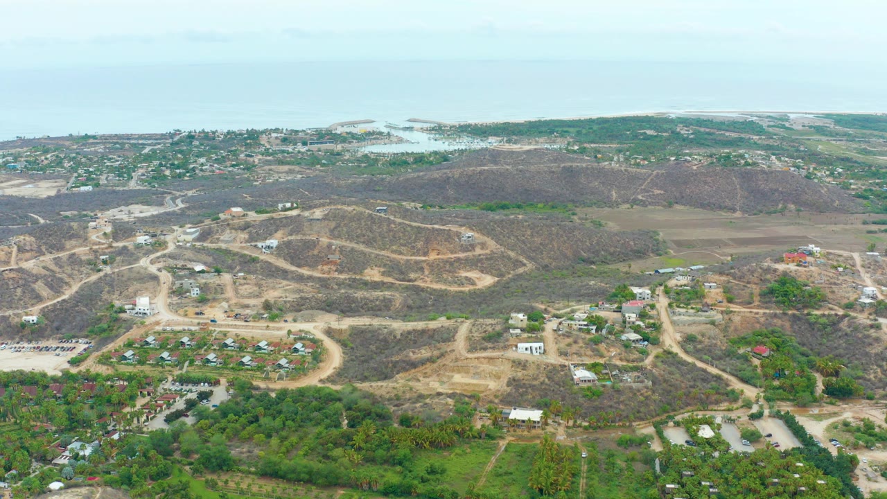 Wide aerial view over La Playita Los Cabos with dry hills, winding dirt roads, scattered buildings, green patches, distant water, and no visible people.
