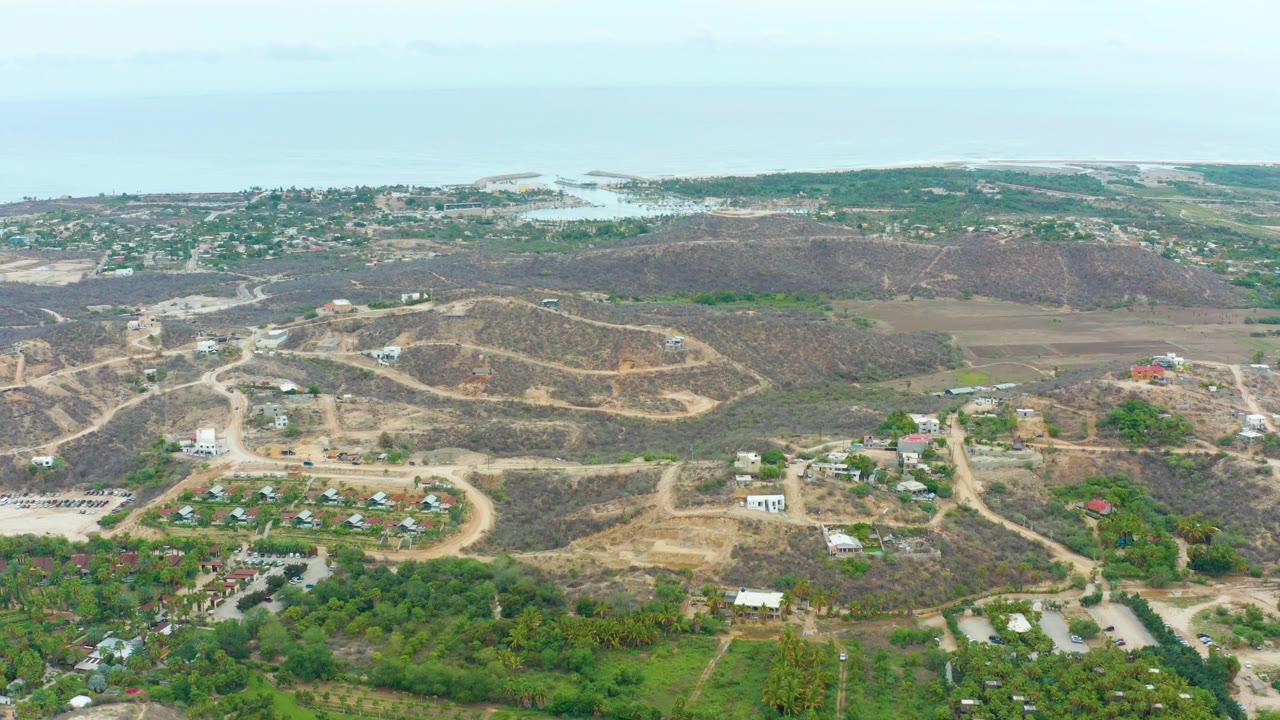 Aerial frame from La Playita Los Cabos showing pale dirt roads across dry slopes, small buildings below, green foreground vegetation, distant water, and no visible people.