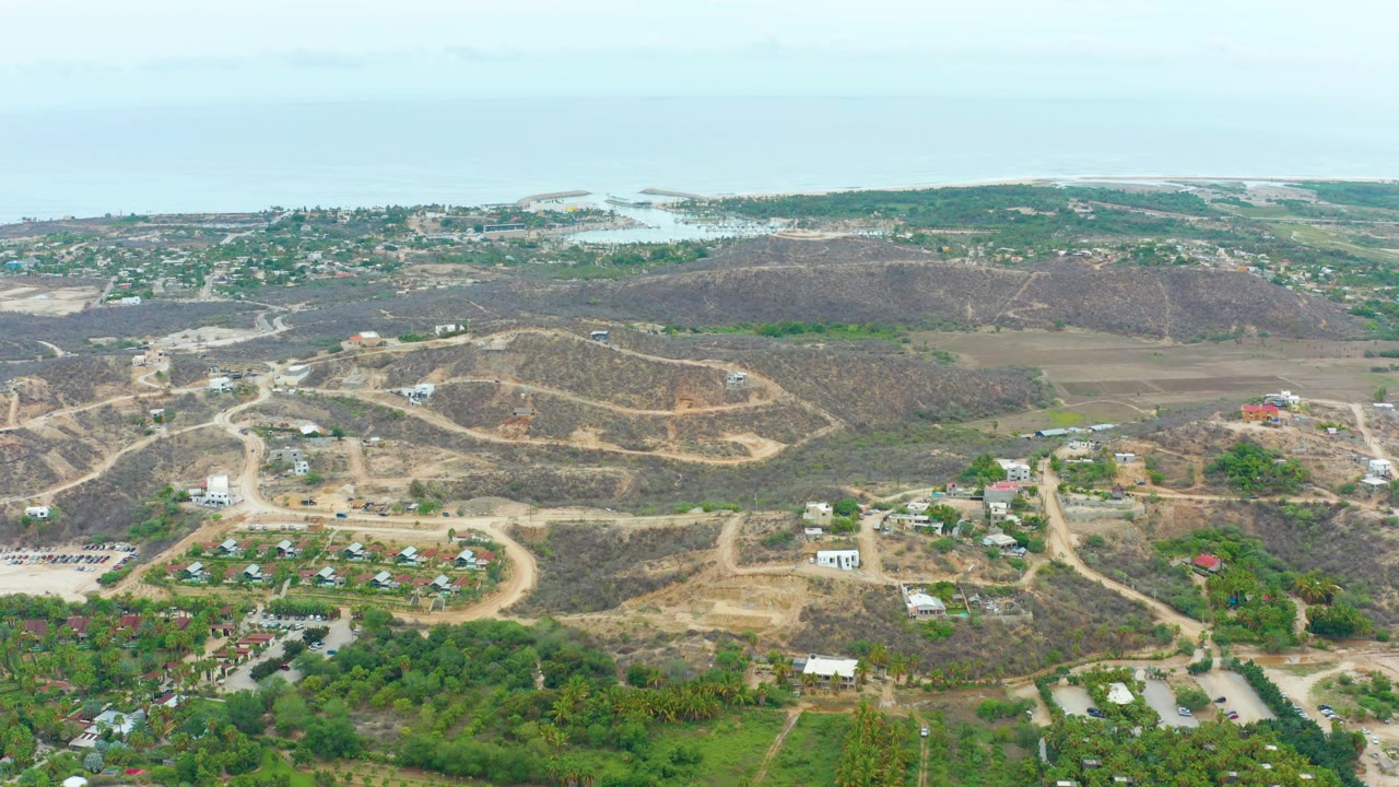 High aerial view in La Playita Los Cabos with dry hillside road patterns, scattered structures, denser green areas in the lower frame, distant water, and no visible people.