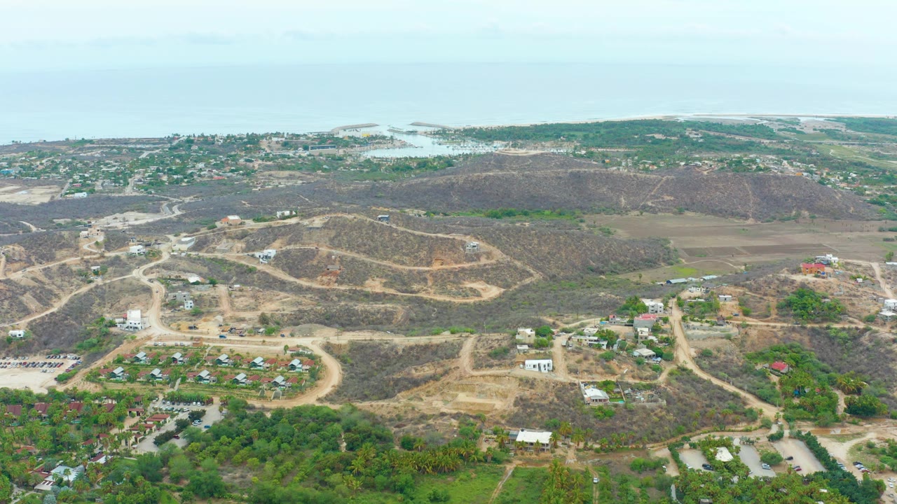 Wide drone view of roads crossing dry hills with buildings and greenery below.
