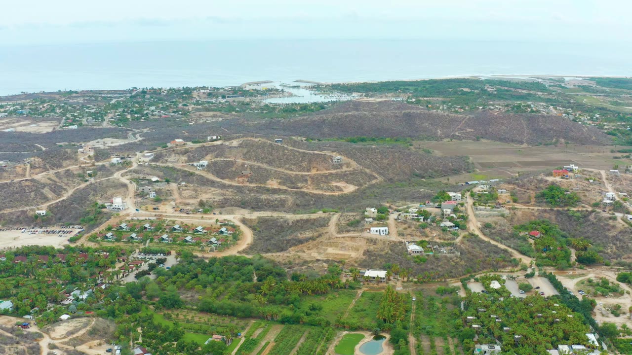 Wide aerial frame over La Playita Los Cabos showing brown hills, winding dirt roads, green fields, scattered buildings, distant homes, pale sky, and no visible people.