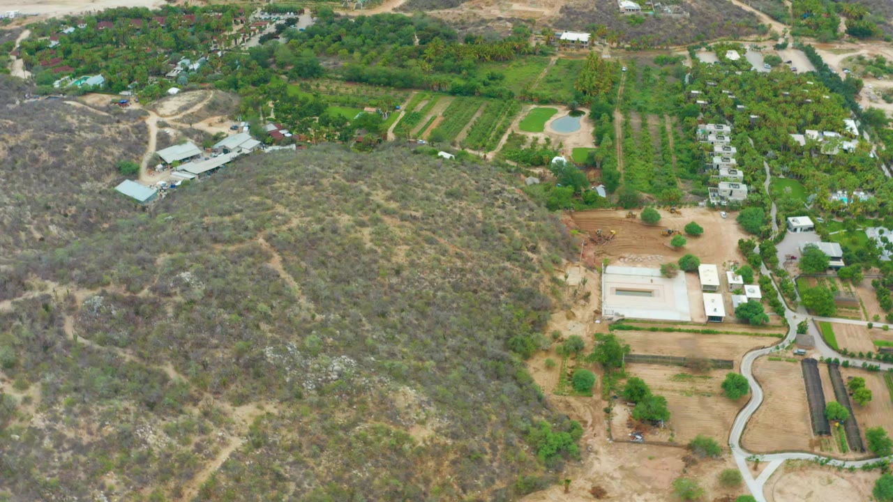 Aerial frame from La Playita Los Cabos showing a dry scrub hill in the foreground, green cultivated rows, farm buildings, dirt roads, and no visible people.