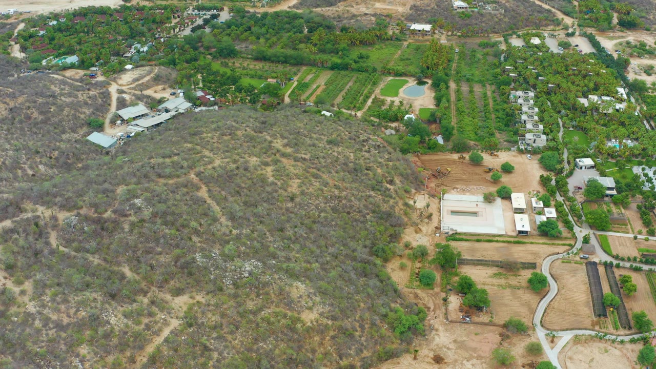 La Playita Los Cabos aerial frame showing cultivated green rows near the top of frame, dry scrubland across the foreground, several light-roofed structures, and no visible people.