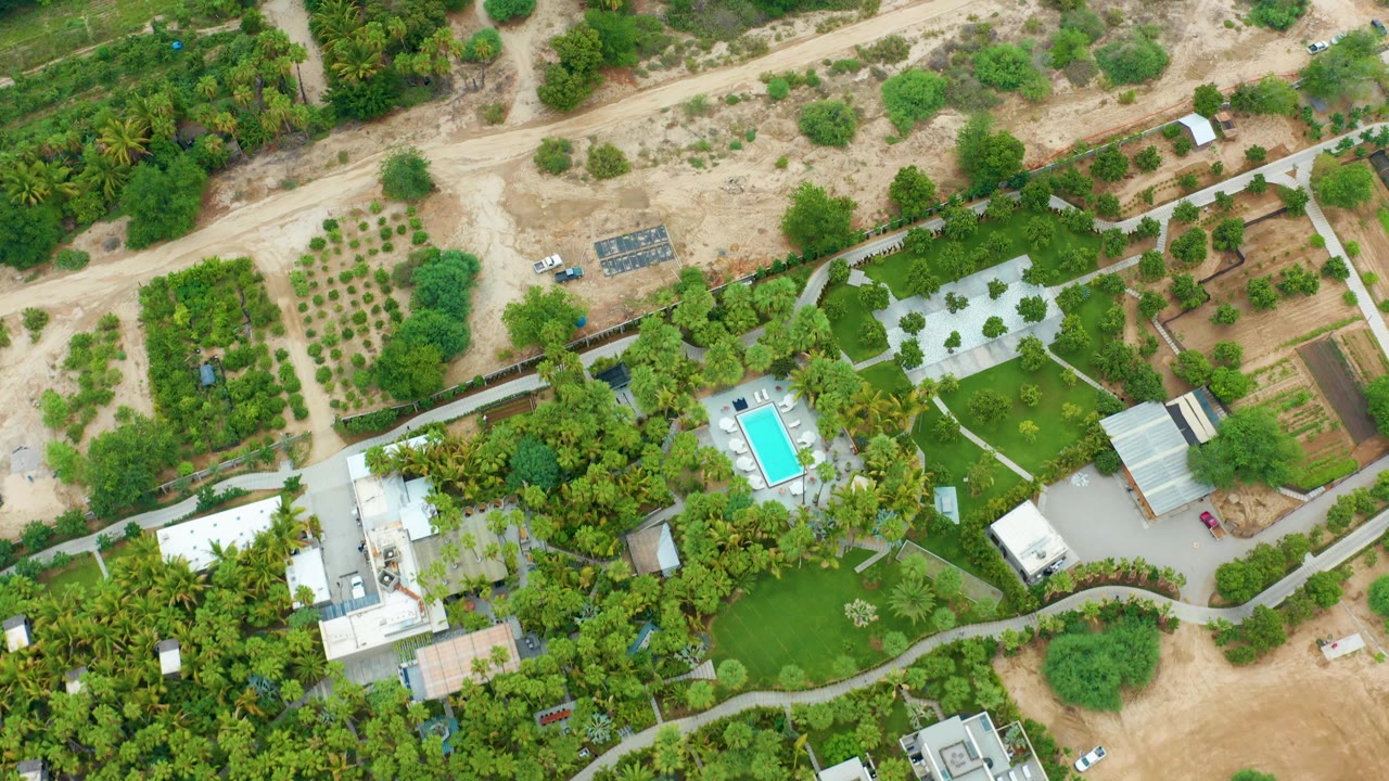 Overhead frame from La Playita Los Cabos showing a white vehicle on a pale dirt road above green residential gardens, roofs, paths, and a swimming pool, with no visible people.