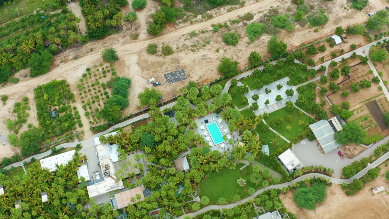 Top-down La Playita Los Cabos frame showing the vehicle near the upper dirt road, with orchards or garden rows, dense greenery, homes, a pool, and paved paths below, with no visible people.