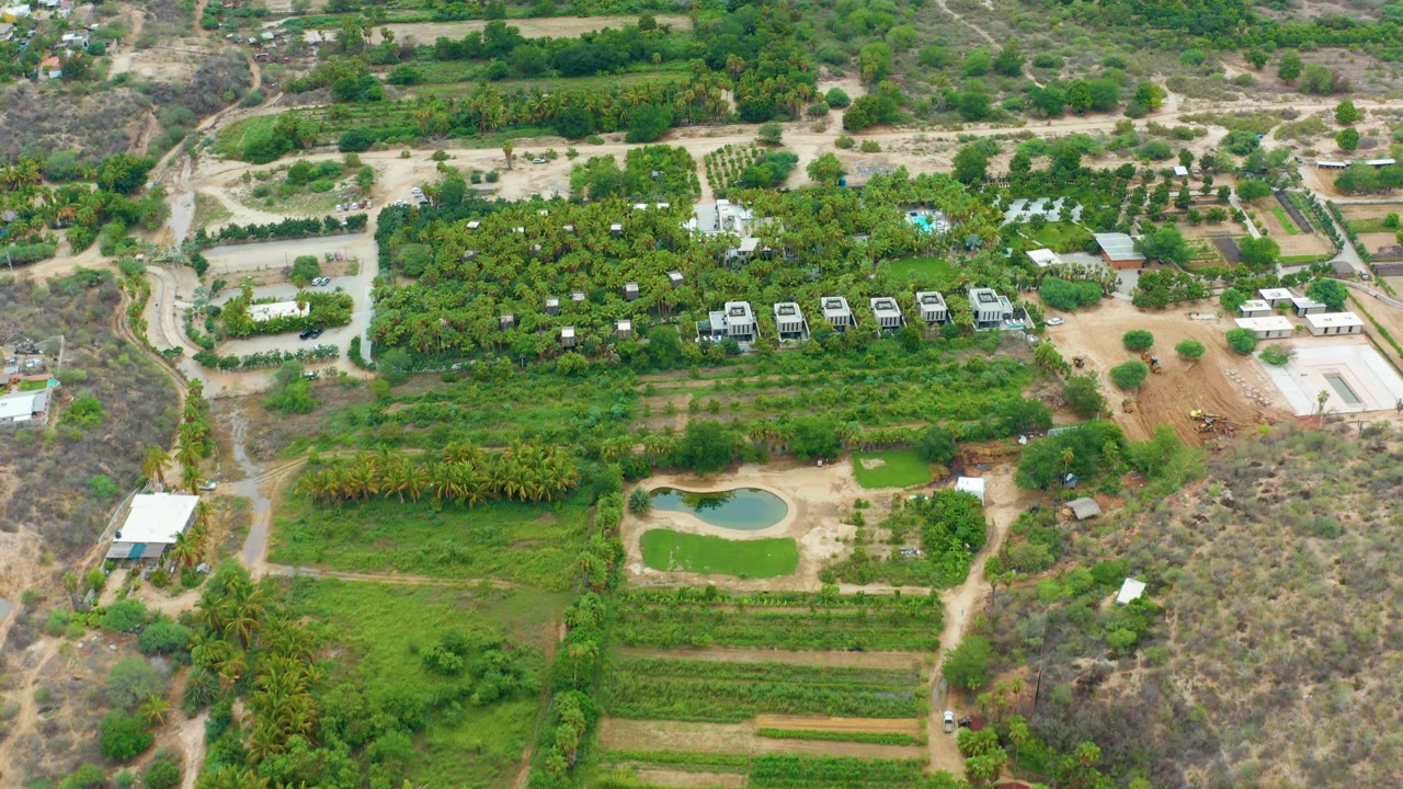 Farm landscape seen from above with crop rows, pond, dirt roads, and white structures.