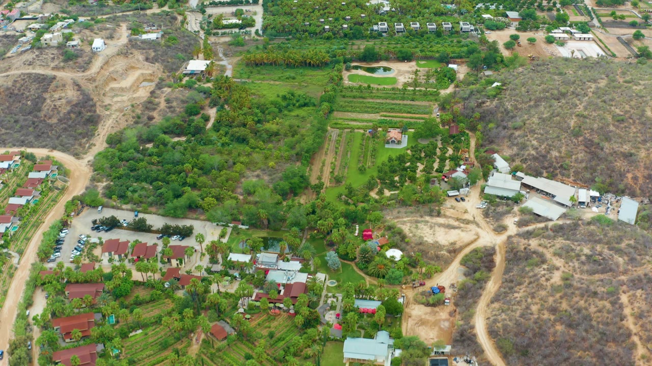 Aerial frame over La Playita Los Cabos showing green farm plots, red-roofed residential buildings, pale dirt roads, dry hillside, and no visible people.