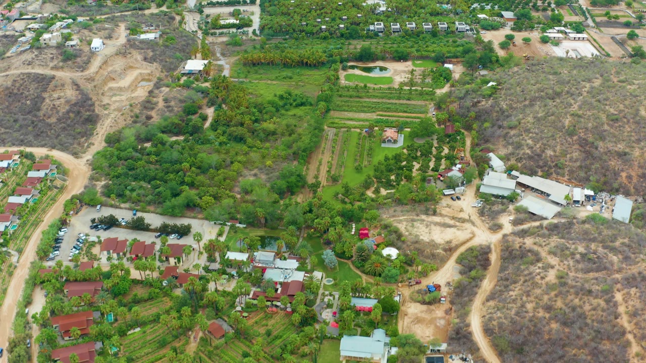 Drone view of farm plots, trees, buildings, and scrub-covered hills.