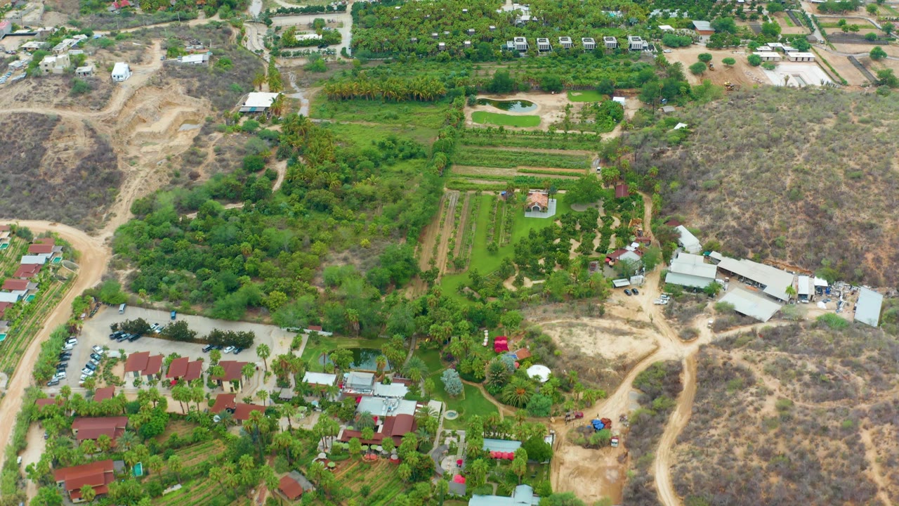 La Playita Los Cabos aerial frame showing a parking area with cars at lower left, red-roofed structures, green fields, dry tracks, hillside brush, and no visible people.