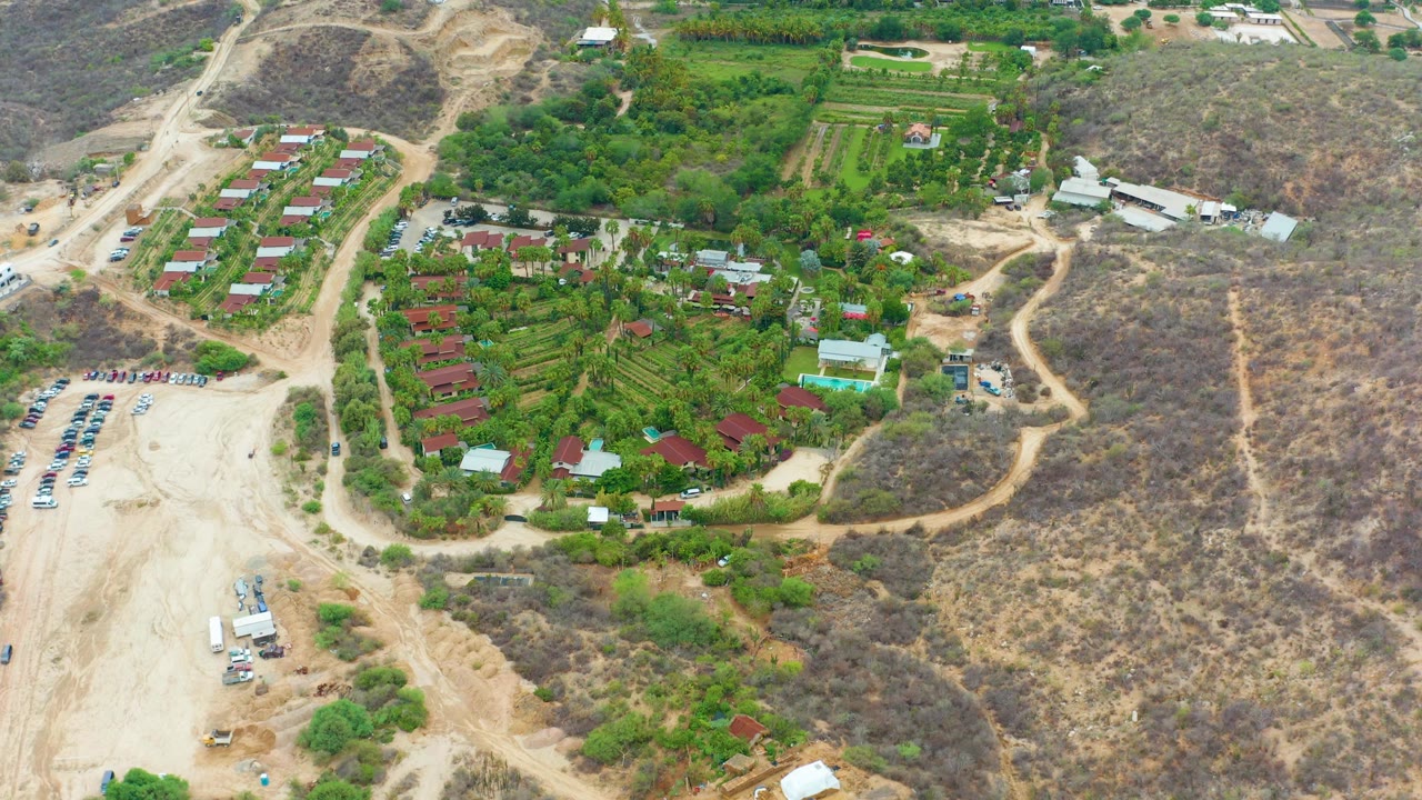 Opening aerial frame over La Playita Los Cabos, Mexico, showing cultivated green plots, red roof buildings, a pale dirt parking area with vehicles, dry hills, and no visible people.