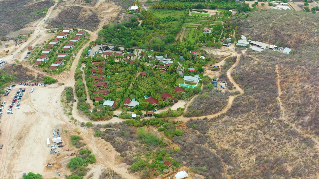 Aerial frame at 0:01 over La Playita Los Cabos, Mexico, with the parking lot lower left, green fields and red roofs centered, dry hillside trails to the right, and no visible people.