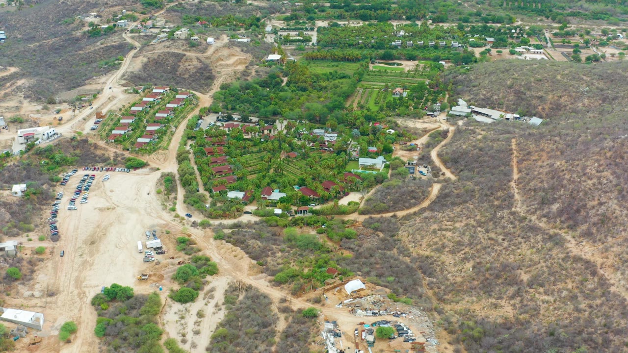 Aerial view in La Playita Los Cabos showing the same dirt-road network, cultivated green area, small buildings, parked vehicles, and scrub-covered hillside with no visible people.