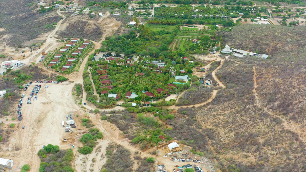 Aerial frame with parking area, dirt road, greenery, and hillside buildings.