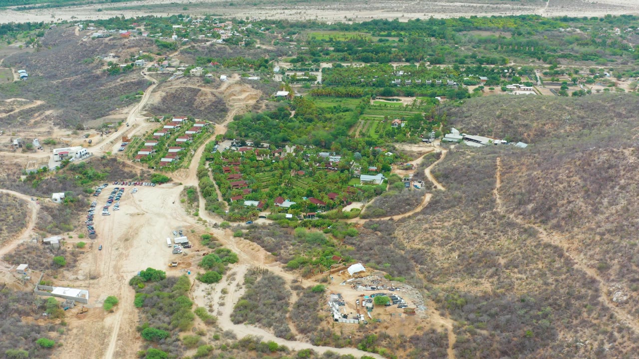 At 0:02, the aerial view holds on La Playita Los Cabos with dry hillside terrain on the right, green planted areas and roofs in the middle, a pale dirt road below, and no visible people.