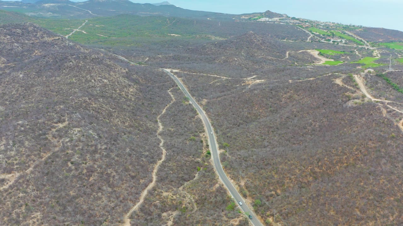 High drone view in La Playita Los Cabos with the winding paved road left of center, dusty footpaths across the hills, green fields in the distance, and no visible people.
