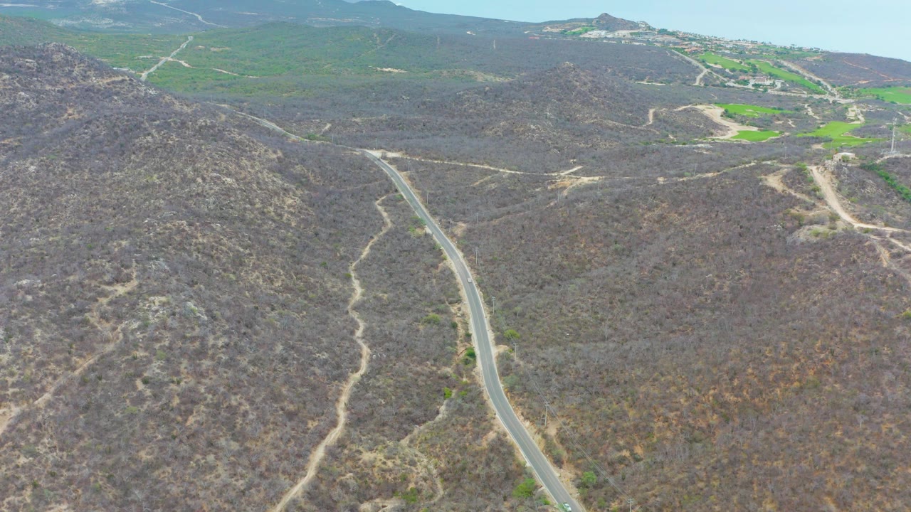 Drone frame from La Playita Los Cabos showing a paved road along the lower left, scrub-covered brown hills across the foreground, bright green fields beyond, and no visible people.