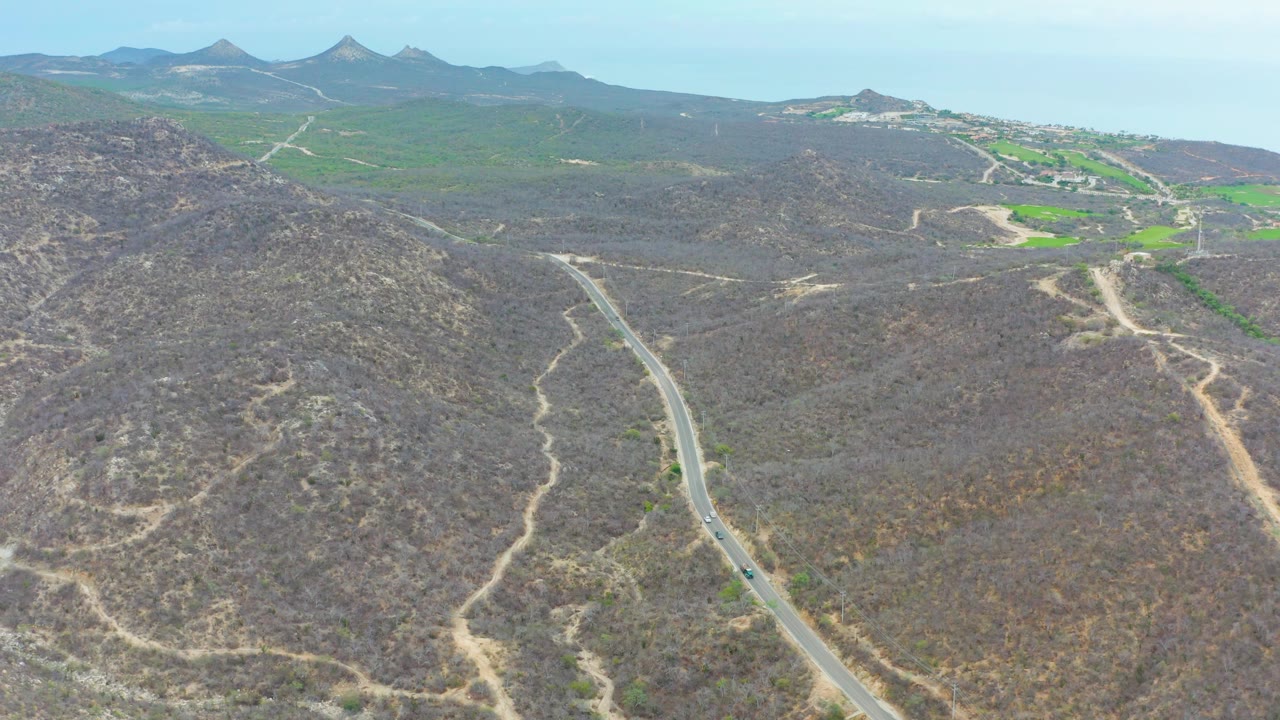 Aerial frame from La Playita Los Cabos showing a winding paved road entering from the lower left through dry brown hills, with green patches and distant mountains beyond, no visible people.