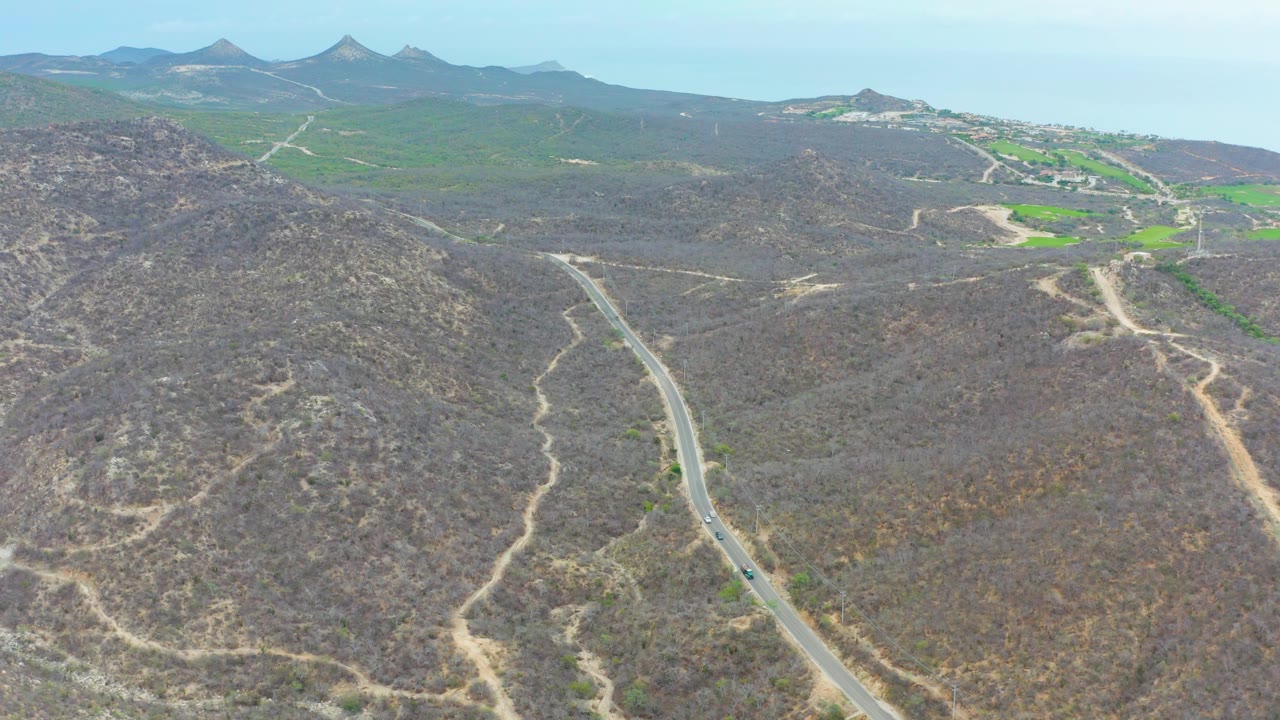 Aerial frame from La Playita Los Cabos with the road curving up from the lower left, pale dirt tracks crossing scrubby hills, and green valley patches near the horizon, no visible people.