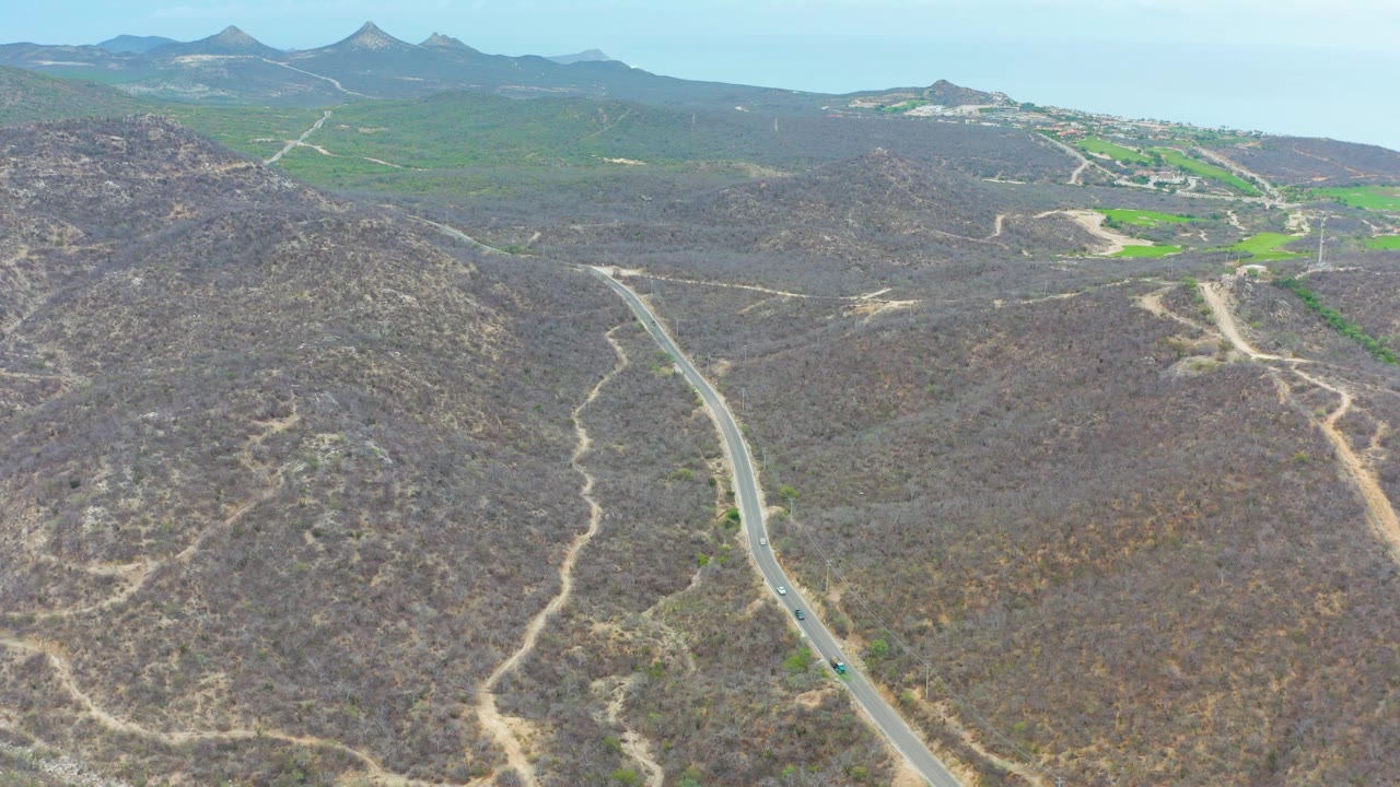 Aerial frame from La Playita Los Cabos showing the paved road bending through the lower left hillside, scattered dirt paths across the center slope, and green fields in the distance, no visible people.