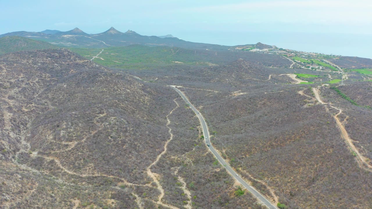 Aerial frame from La Playita Los Cabos showing a pale road curving through brown scrub hills toward distant mountains and open blue sky, with no visible people.