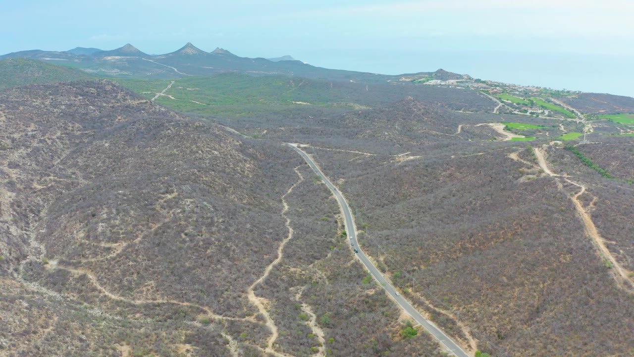 The La Playita Los Cabos road scene shows the pavement cutting through scrubby hills, with dirt tracks, bright green areas, distant buildings, mountains, sky, and no visible people.