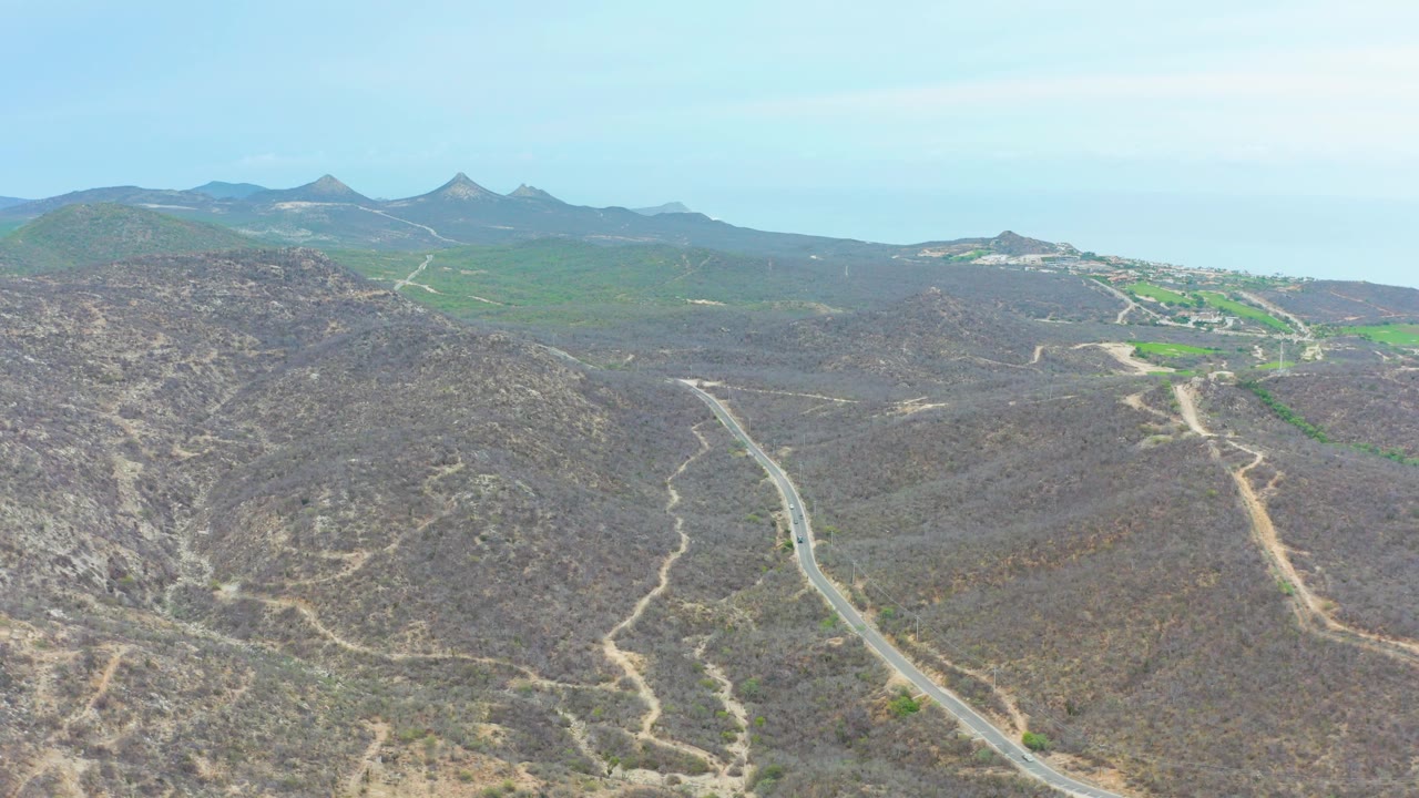 Opening aerial frame over La Playita Los Cabos, Mexico, with dry scrub hills in front, a paved road curving through the valley, green land in the distance, and no visible people.