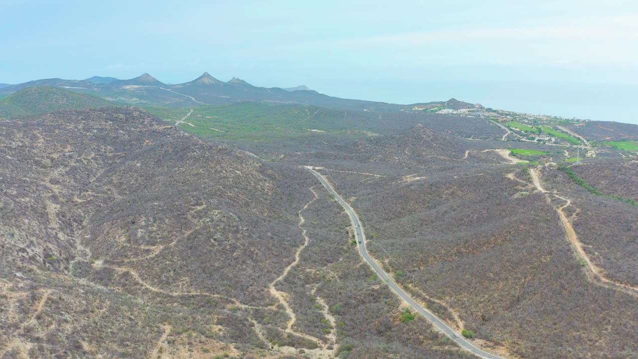 Aerial frame over La Playita Los Cabos, Mexico, with brown hills and dirt routes in the foreground, green terrain and a small developed area near the horizon, and no visible people.