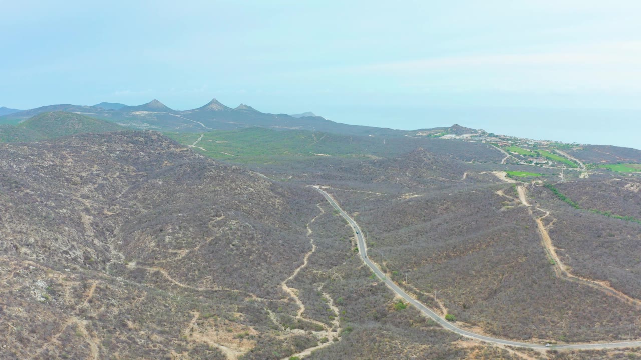 At one second, the aerial view holds on the curved road through La Playita Los Cabos hills, with brown scrubland in the foreground, green fields beyond, and no visible people.