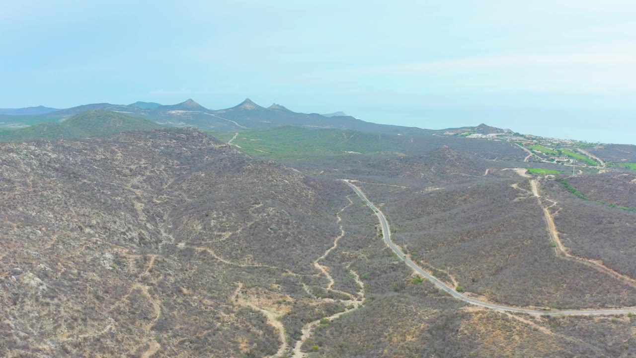 Aerial view in La Playita Los Cabos with a curving road across scrubby hills, green fields in the distance, pointed mountain ridges, and no visible people.