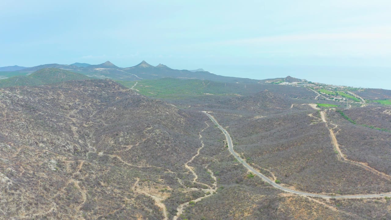 Aerial frame in La Playita Los Cabos with a road bending through the center of dry hills, faint side trails, distant green areas, mountain silhouettes, and no visible people.