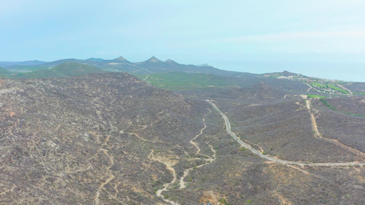 Drone view in La Playita Los Cabos looking across rocky brown hills, branching dirt roads, green distant ridges, clear blue sky, and no visible people.