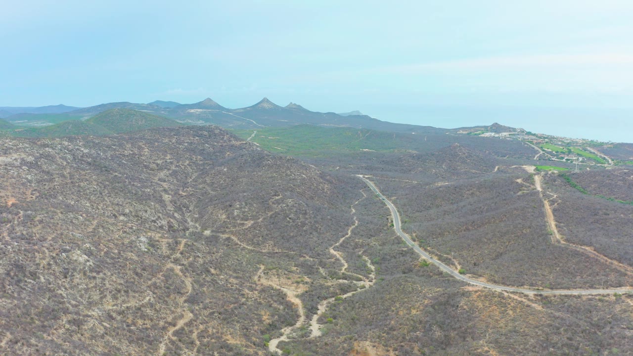 Aerial landscape in La Playita Los Cabos with a winding road descending through dry hills, rocky foreground texture, distant ridges, pale sky, and no visible people.