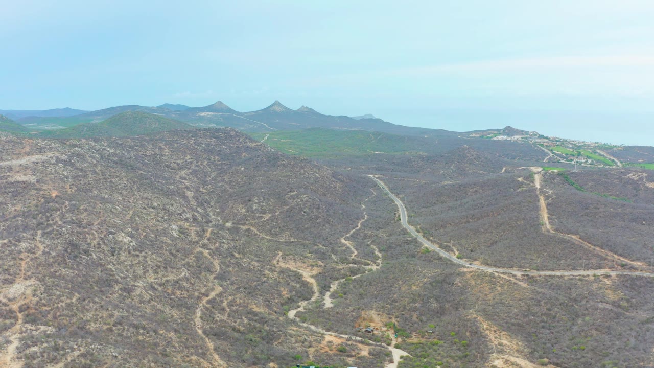 A wide aerial view over La Playita Los Cabos shows a winding dirt road crossing dry rocky hills, green distant slopes, a pale blue sky, and no visible people.