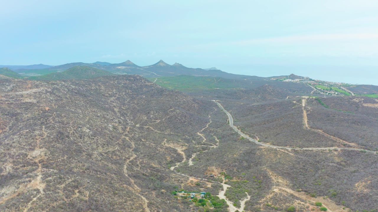 Opening aerial frame over La Playita Los Cabos dry hills, with pale dirt roads crossing brown scrubland, distant green ridges, blue sky, and no visible people.