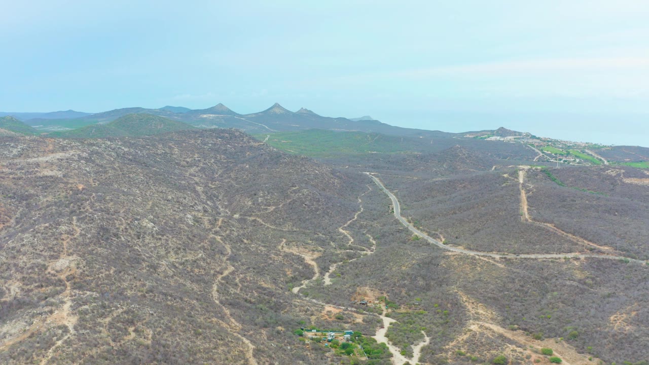 Aerial frame in La Playita Los Cabos showing intersecting dirt tracks through brown hills, green terrain on the horizon, small structures near the lower edge, and no visible people.