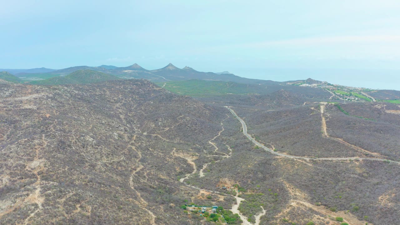 La Playita Los Cabos aerial frame with rugged scrub-covered hills in the foreground, a pale road curving across the midground, distant ridgelines, blue sky, and no visible people.
