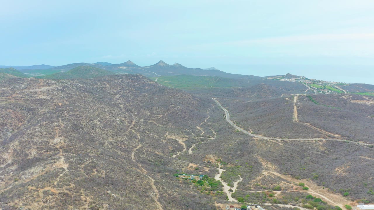 Aerial frame in La Playita Los Cabos, Mexico, showing branching dirt tracks across brown hills, green vegetation near the lower edge, distant ridges, and no visible people.