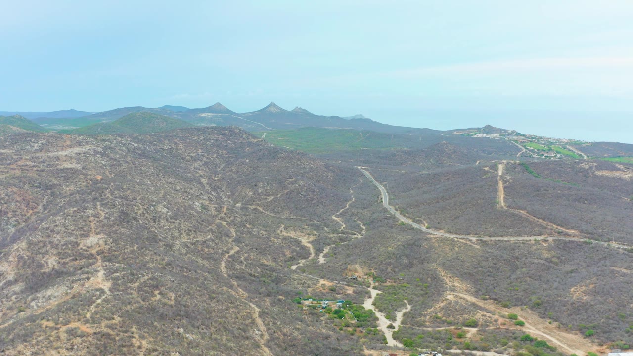 Wide aerial view over La Playita Los Cabos, Mexico, with winding dirt roads in dry terrain, pointed mountain peaks on the horizon, pale sky, and no visible people.