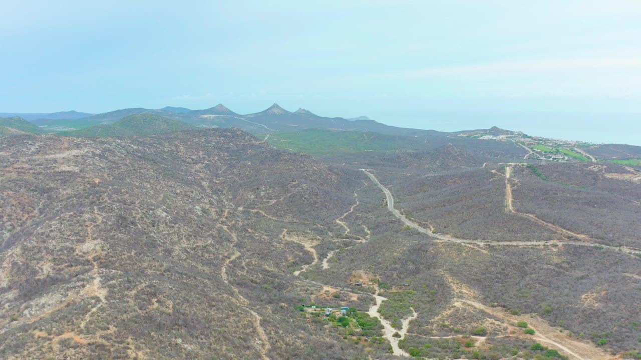 Aerial frame from La Playita Los Cabos, Mexico, with a dirt road leading through arid hills toward distant mountains, small green areas, and no visible people.