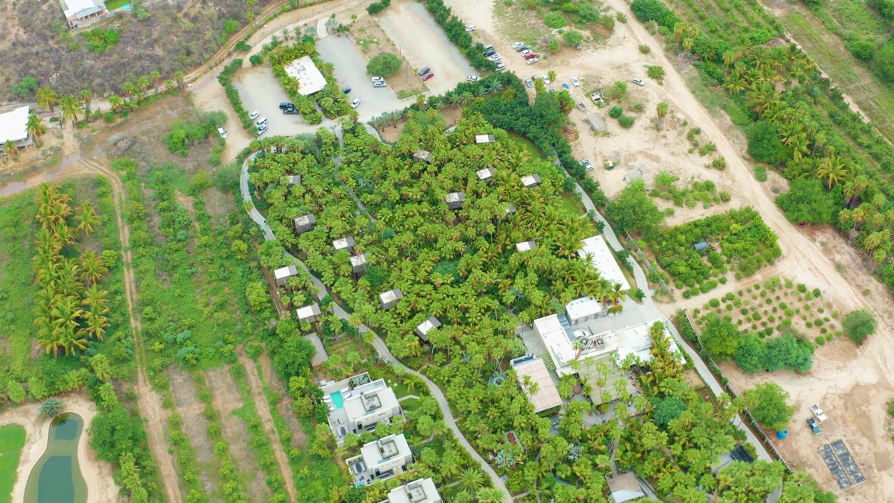 Drone-style overhead frame in La Playita Los Cabos showing the central green property bordered by dirt roads, fields, scattered buildings, vehicles, and no visible people.