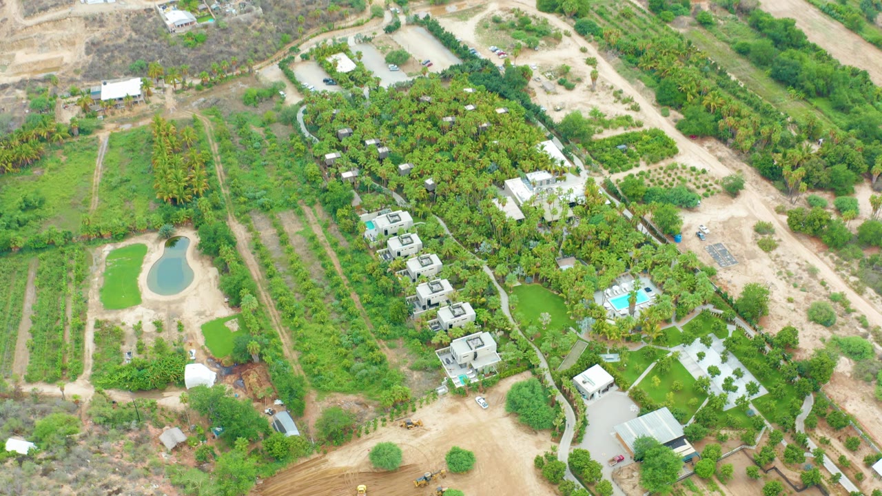 Aerial frame over La Playita Los Cabos farmland shows a small pond at left, planted rows, dirt paths, scattered buildings, dense vegetation, and a blue pool, with no visible people.