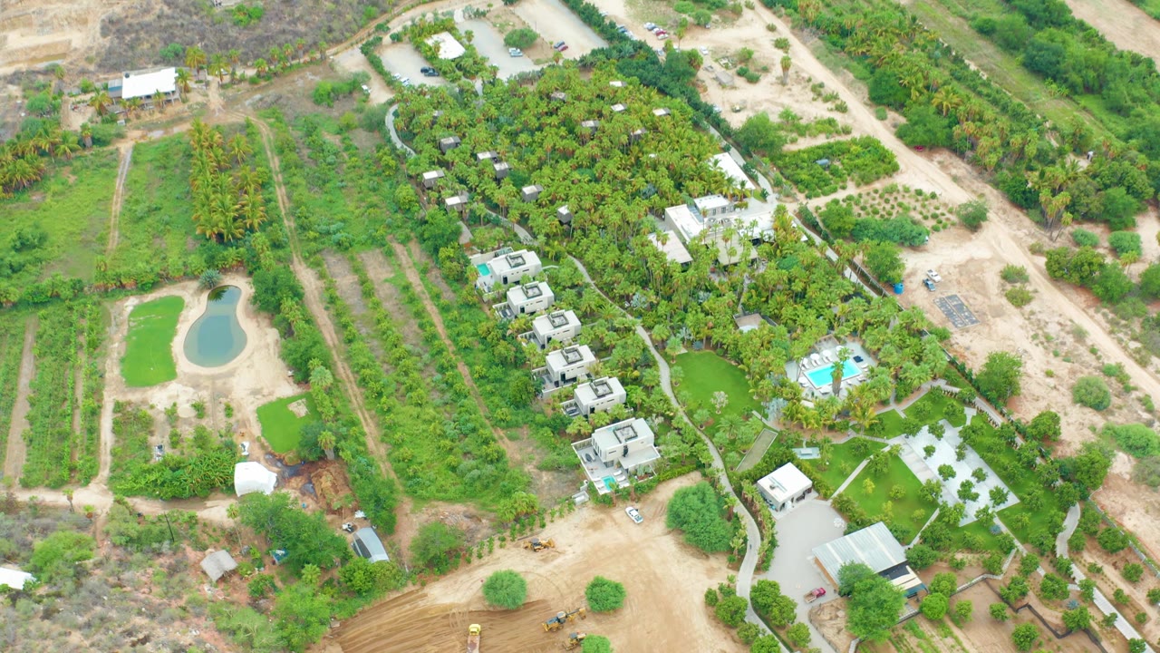 At 0:02, the La Playita Los Cabos aerial frame shows the pond near the left edge, parallel planting rows, a central line of buildings, and a pool surrounded by bright green landscaping, with no visible people.