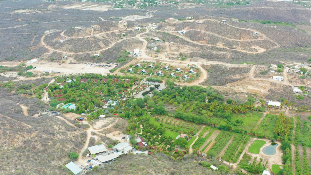 One second into the La Playita Los Cabos aerial view, winding dirt roads cross dry hills above dense green trees, cultivated rows, scattered structures, and no visible people.