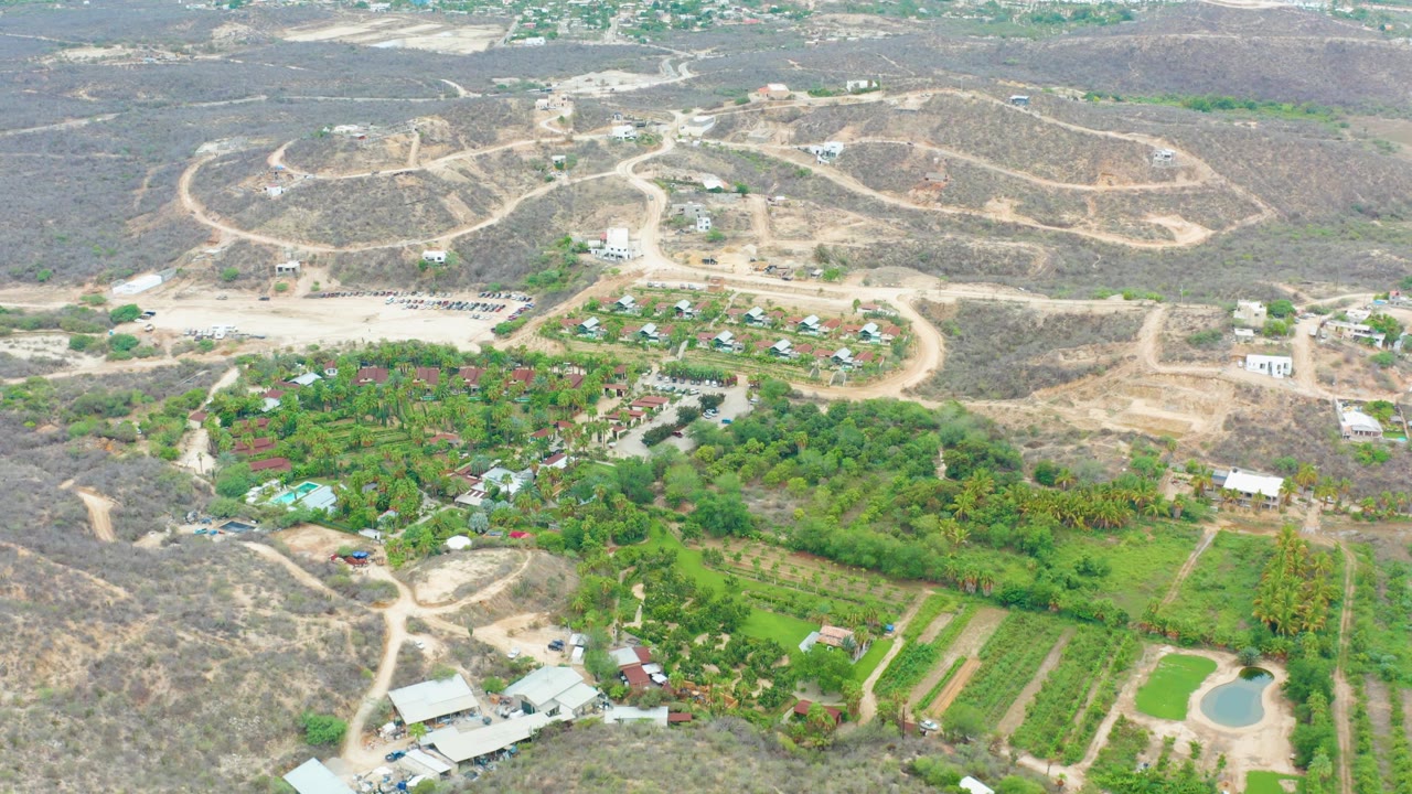 The La Playita Los Cabos frame shows the drone vantage over arid hills, pale road cuts, bright green farm plots, small buildings, a pond at lower right, and no visible people.