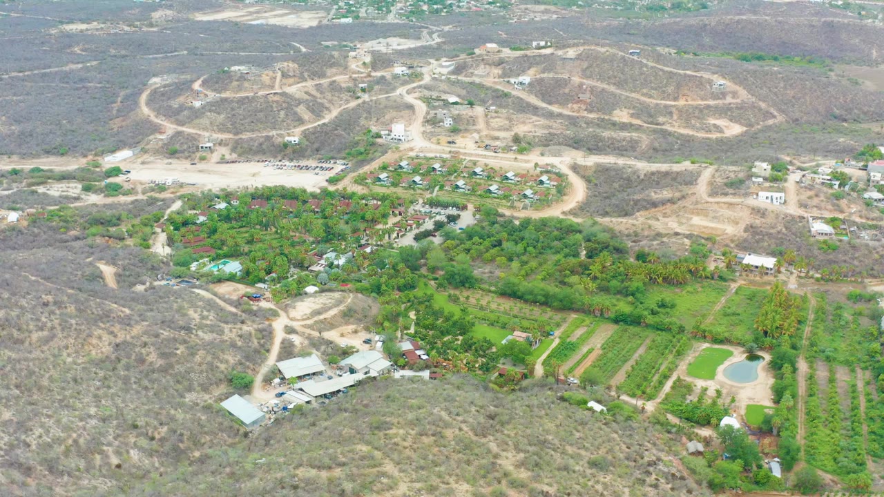 Aerial view over La Playita Los Cabos with dry brown hills above a green cultivated area, scattered buildings, a small pond, and no visible people.