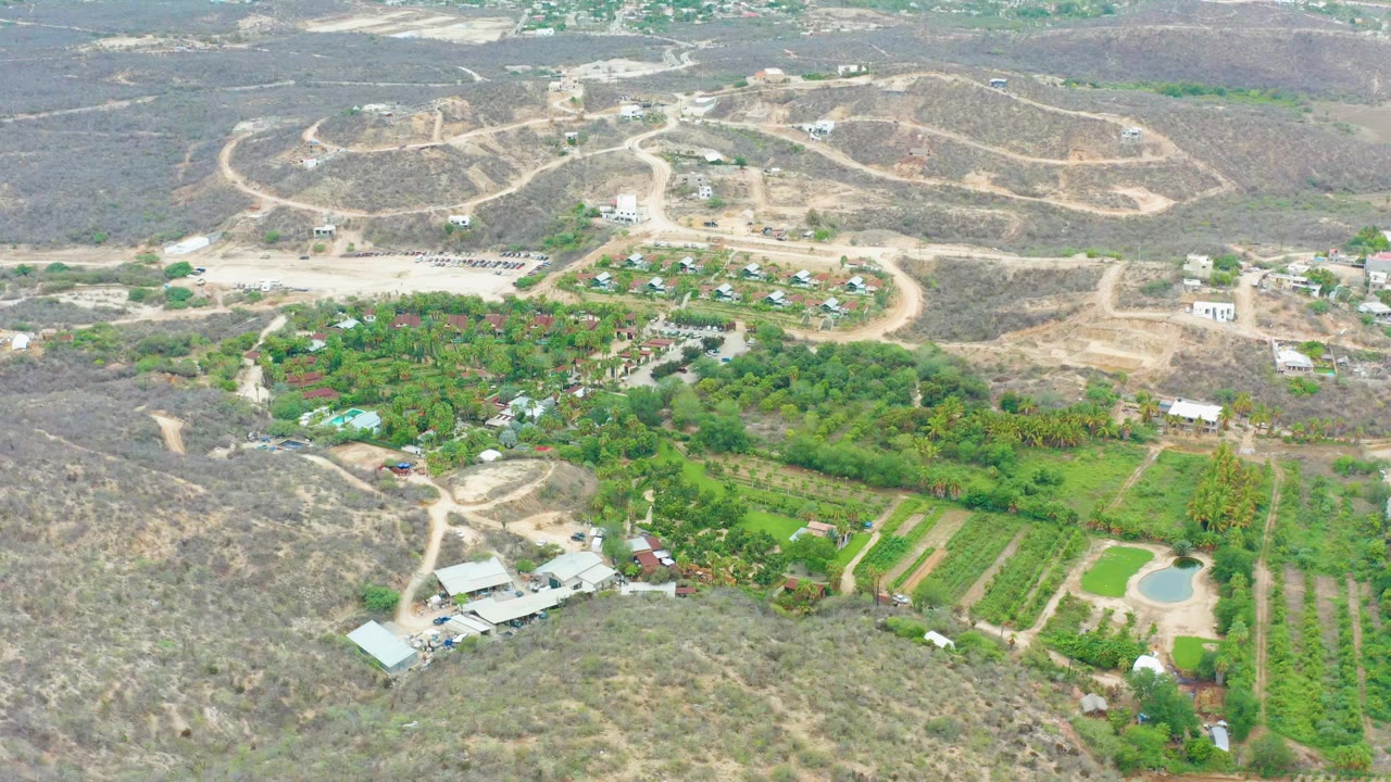 High aerial frame in La Playita Los Cabos showing winding dirt roads through dry terrain, a green grove and fields below, small structures, and no visible people.