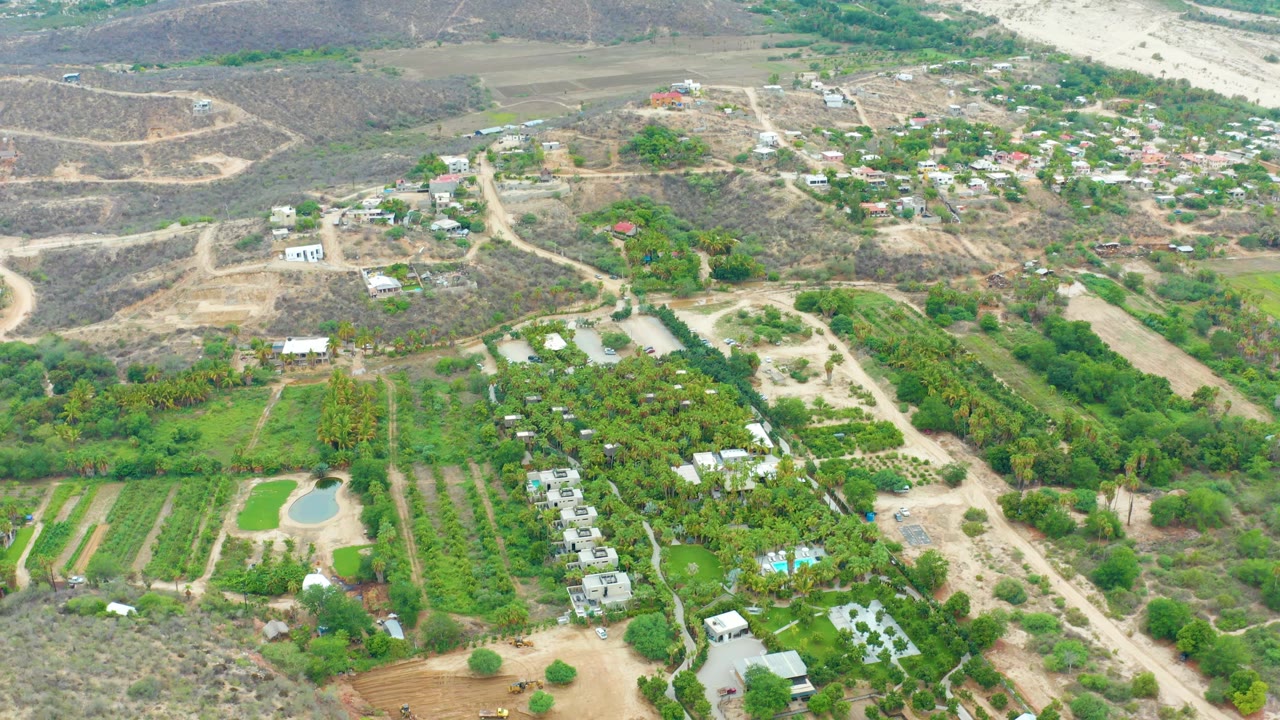 Opening aerial frame over La Playita Los Cabos with green fields in the foreground, scattered small structures, a pale pool, dry hills, and no visible people.