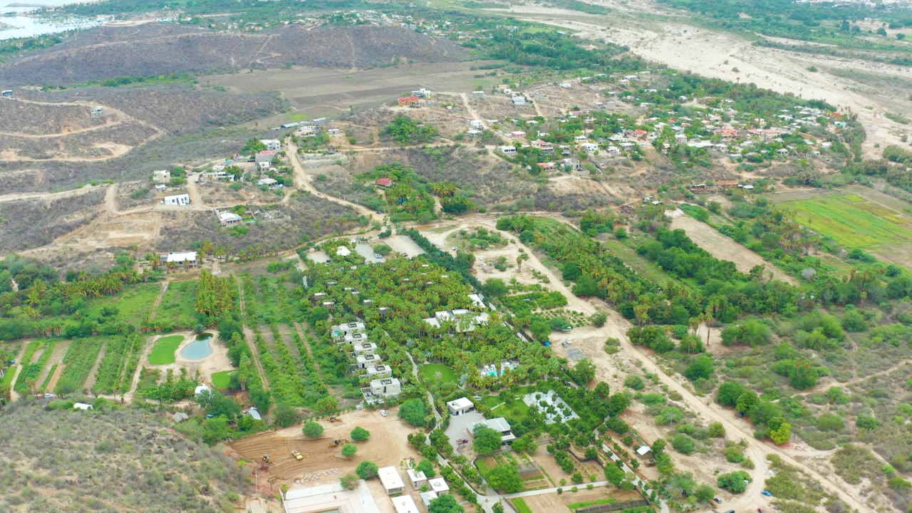 Aerial frame over La Playita Los Cabos showing cultivated green plots in the foreground, scattered houses beyond them, dry hills, a pale riverbed, and no visible people.