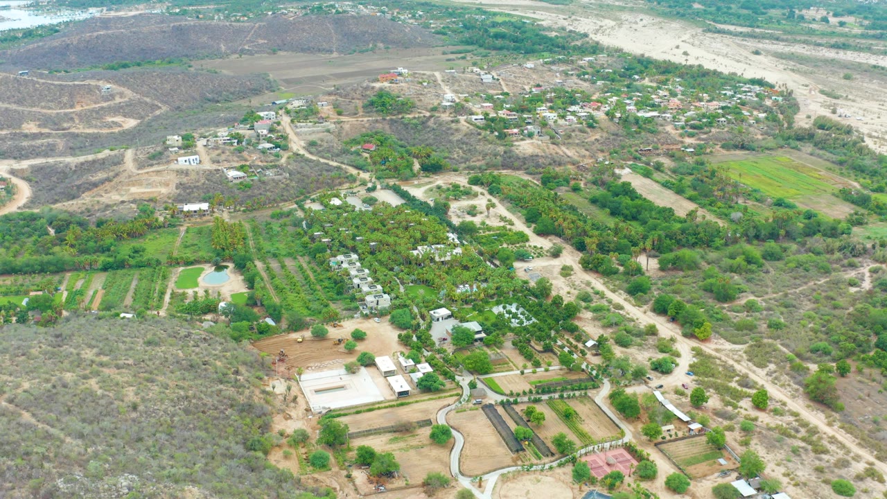 Aerial frame over La Playita Los Cabos showing cultivated green plots, dirt roads, scattered buildings, and dry hillside terrain with no visible people.