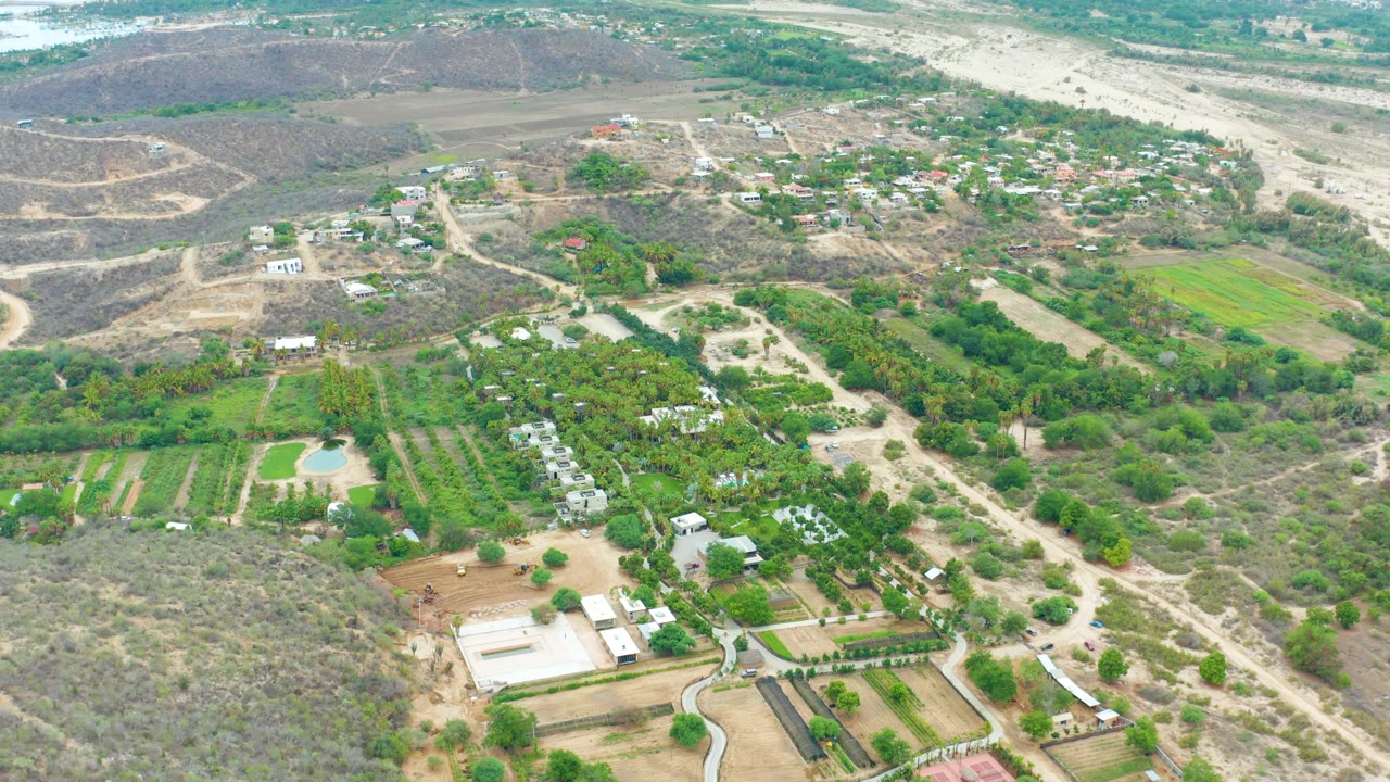 Aerial landscape with farm buildings, planted rows, dirt roads, and dry brushland.